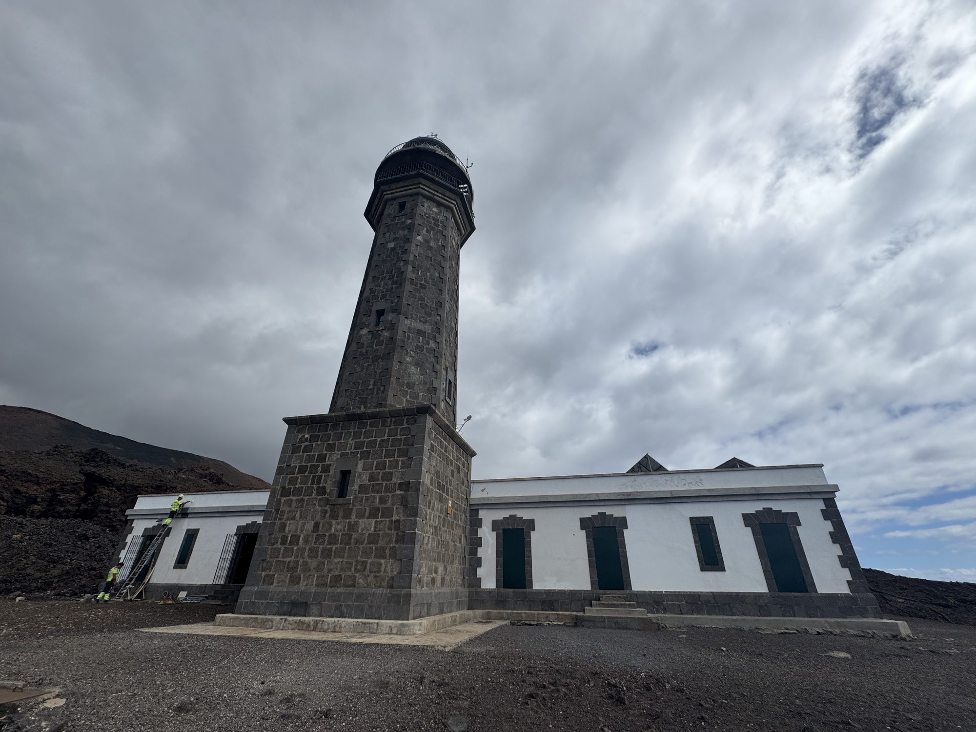 Dark stone tower of the Orchilla lighthouse and its white keeper’s house under a cloudy sky, El Hierro