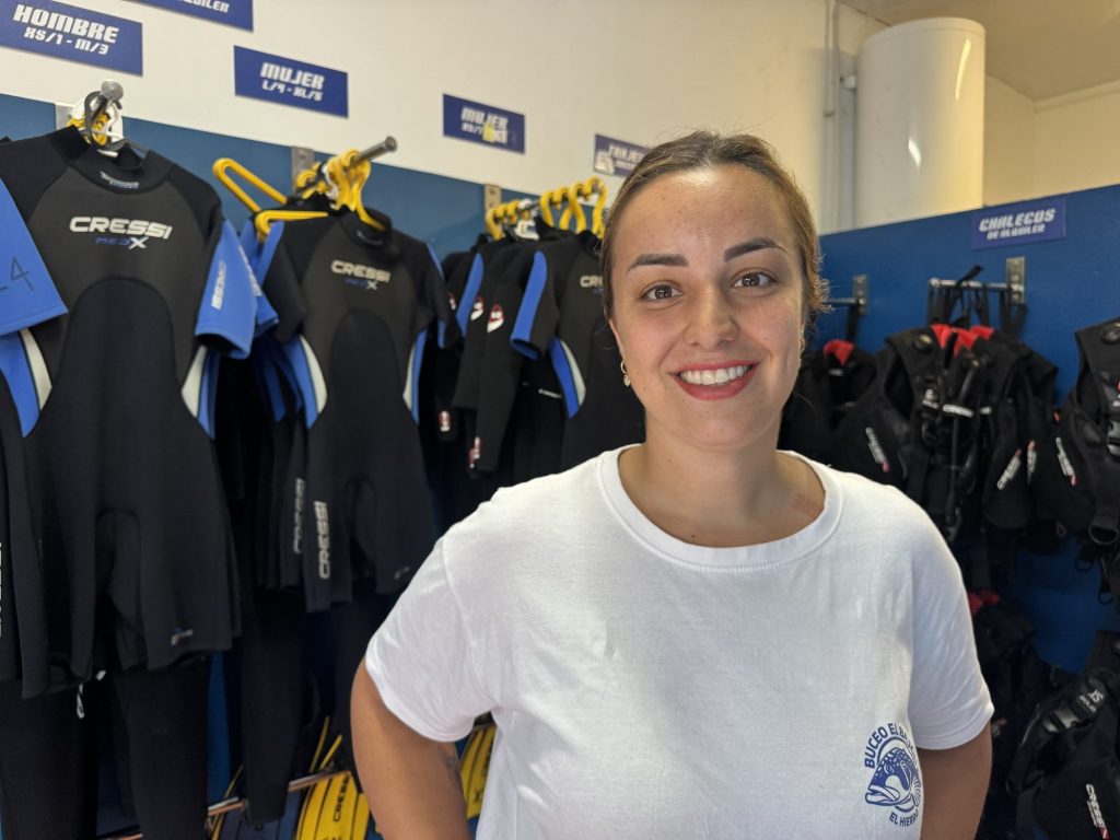 Gisella Horn smiling in front of rows of wetsuits at the Buceo El Bajón dive centre, La Restinga